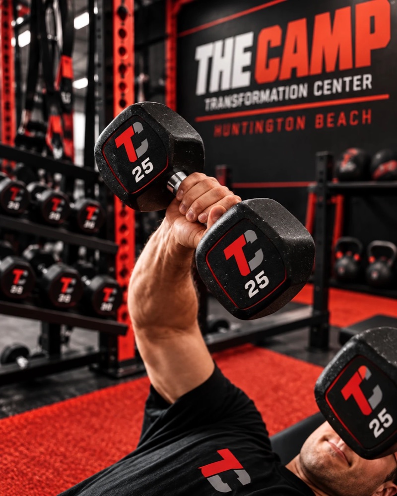 A member pressing Camp-branded dumbbells from a bench.